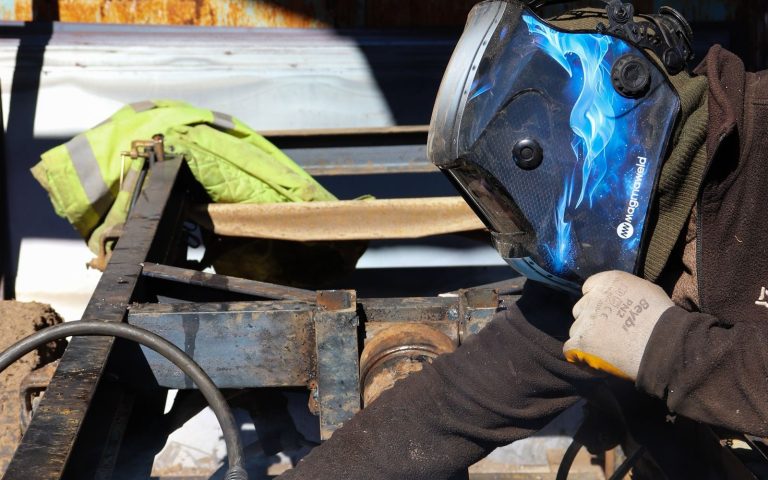 Mobile welder in a helmet welding a steel trailer frame on site outdoors