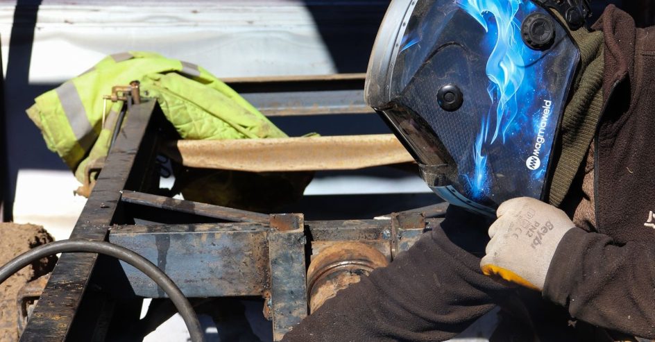 Mobile welder in a helmet welding a steel trailer frame on site outdoors