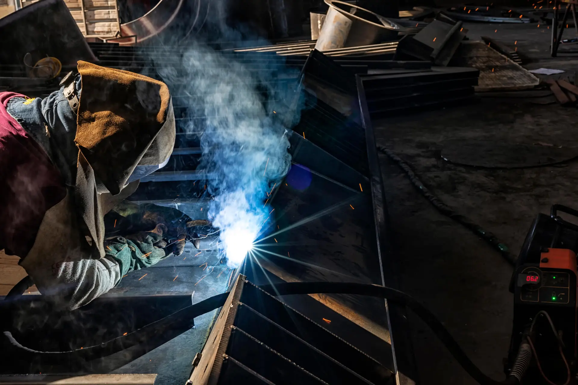 Welder in a leather jacket and helmet welding a steel frame in a dark workshop as smoke rises