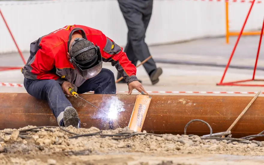 Welder in a red jacket stick-welding a large steel pipe at an outdoor work site
