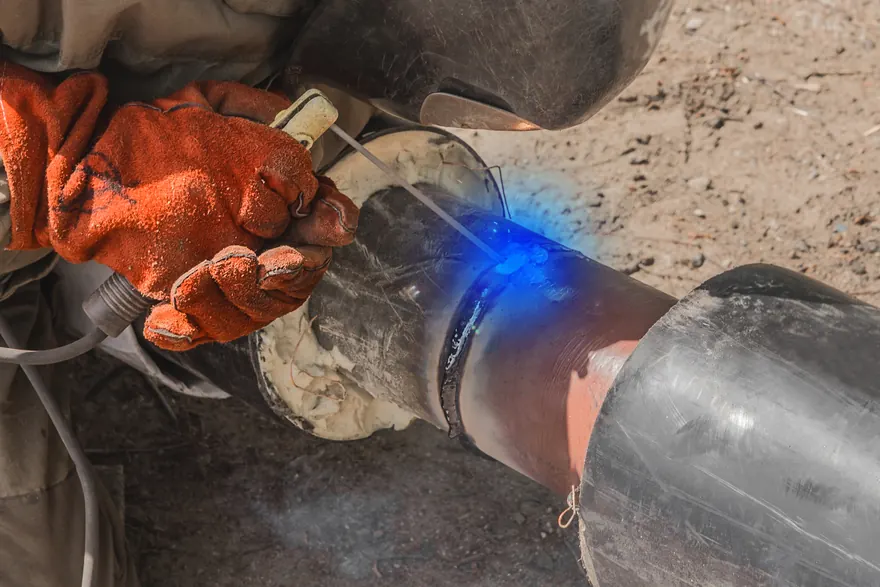 Gloved hands holding an electrode holder as a blue welding arc joins two pipe sections