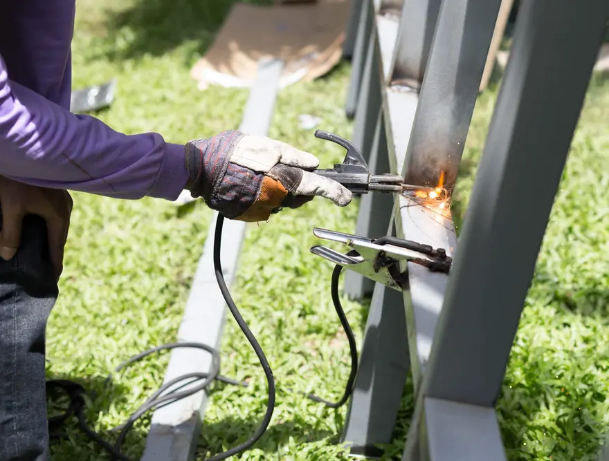 Gloved hand welding a steel frame laid across sawhorses on a lawn
