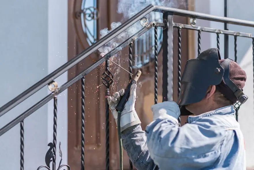 Welder in a helmet welding a wrought iron stair railing at a front entrance