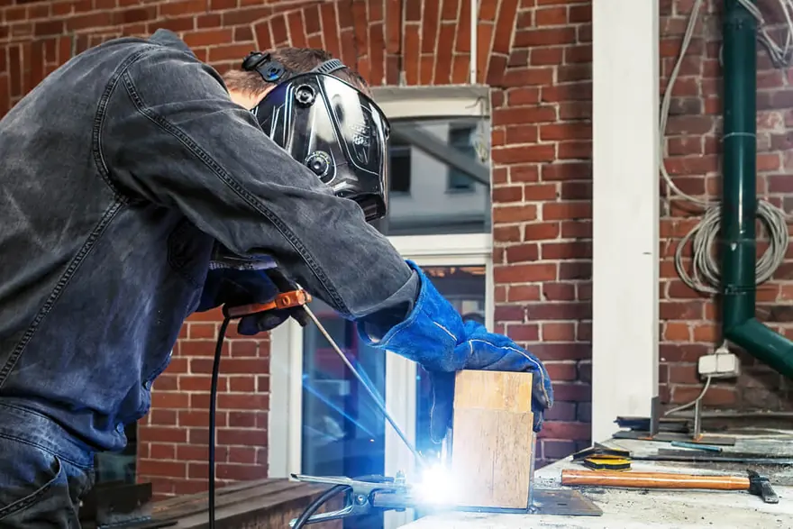 Welder in a helmet working at a bench beside a window in a brick building